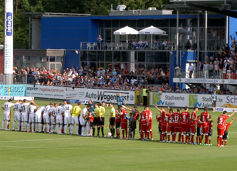  Dann konnte es losgehen: Die beiden Teams grüßten ihre Fans. Im Hintergrund das eigenwillige, dennoch charmante ViP-Gebäude.